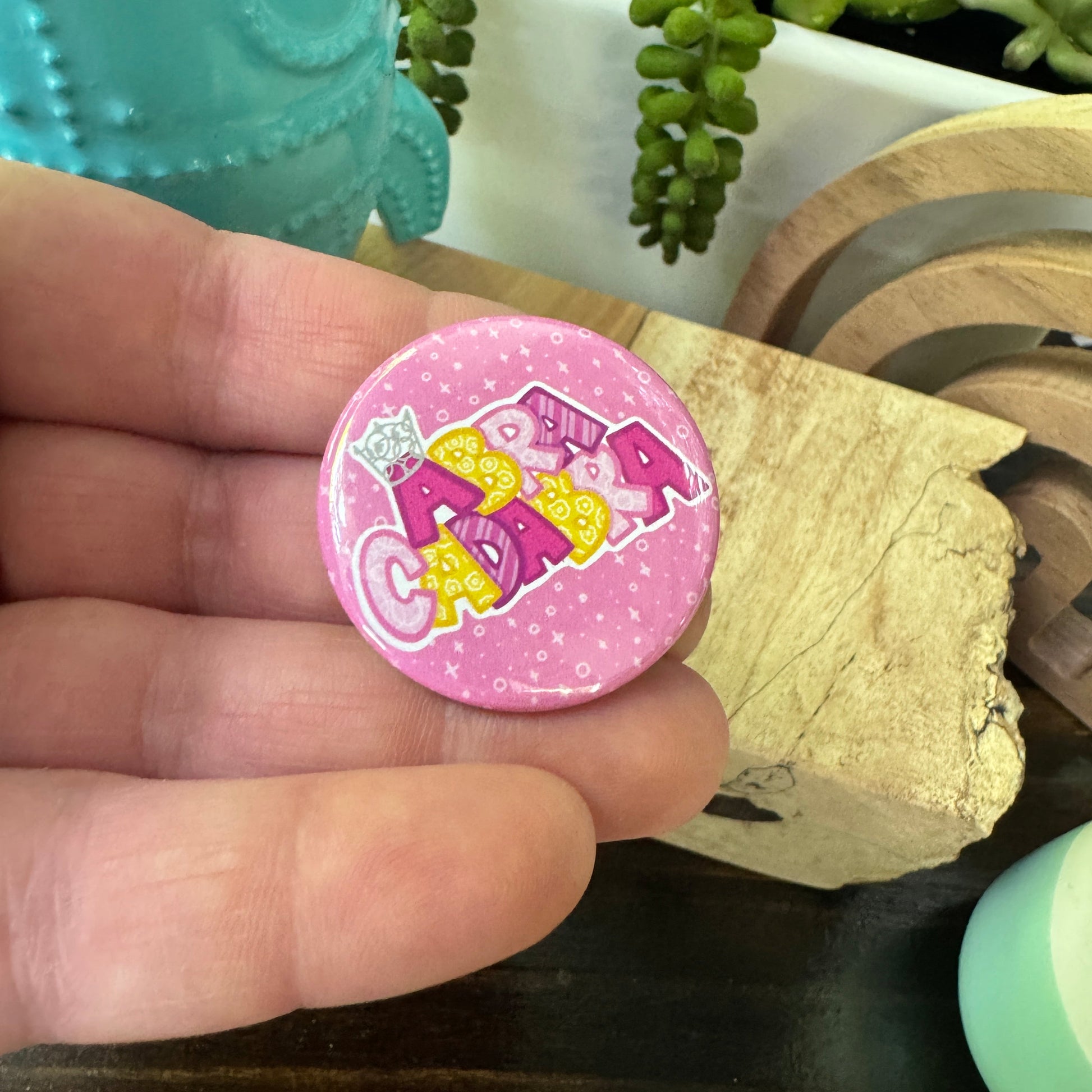 Pink button with 'Abracadabra' text held between fingers, surrounded by decorative items.