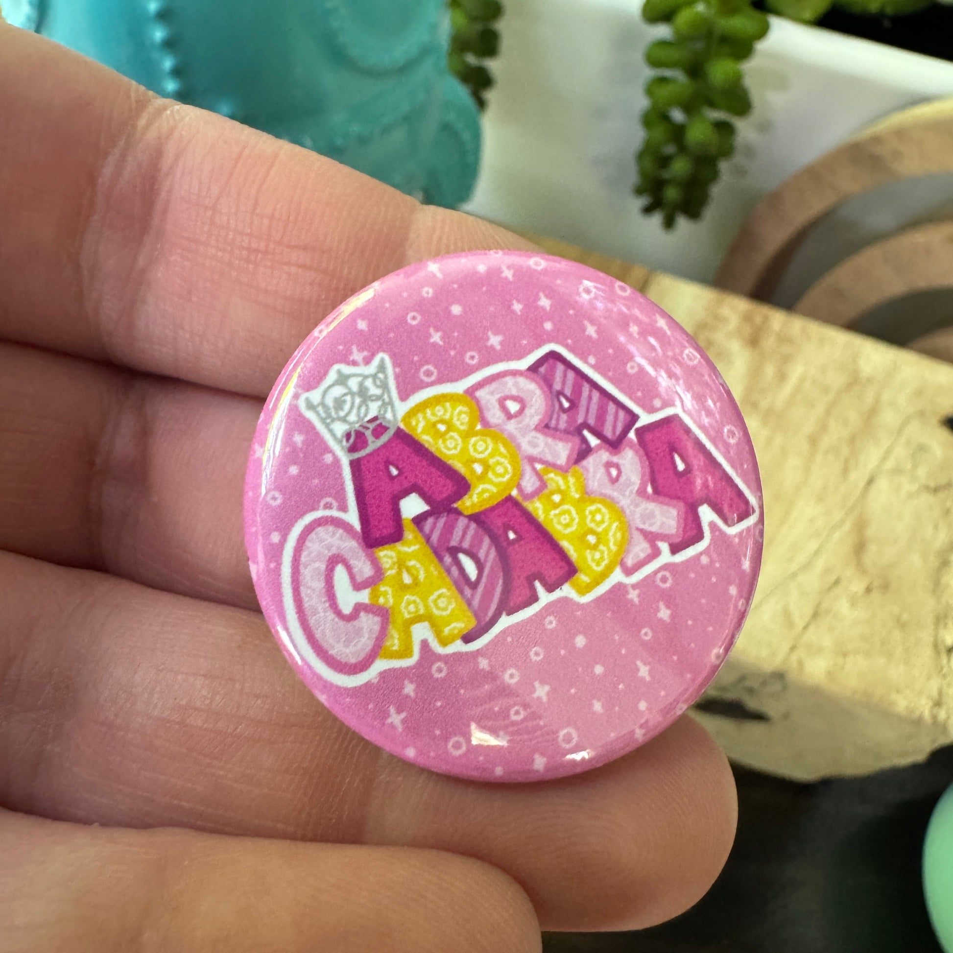 Pink button held in a hand with yellow and pink text held over some decorative items.