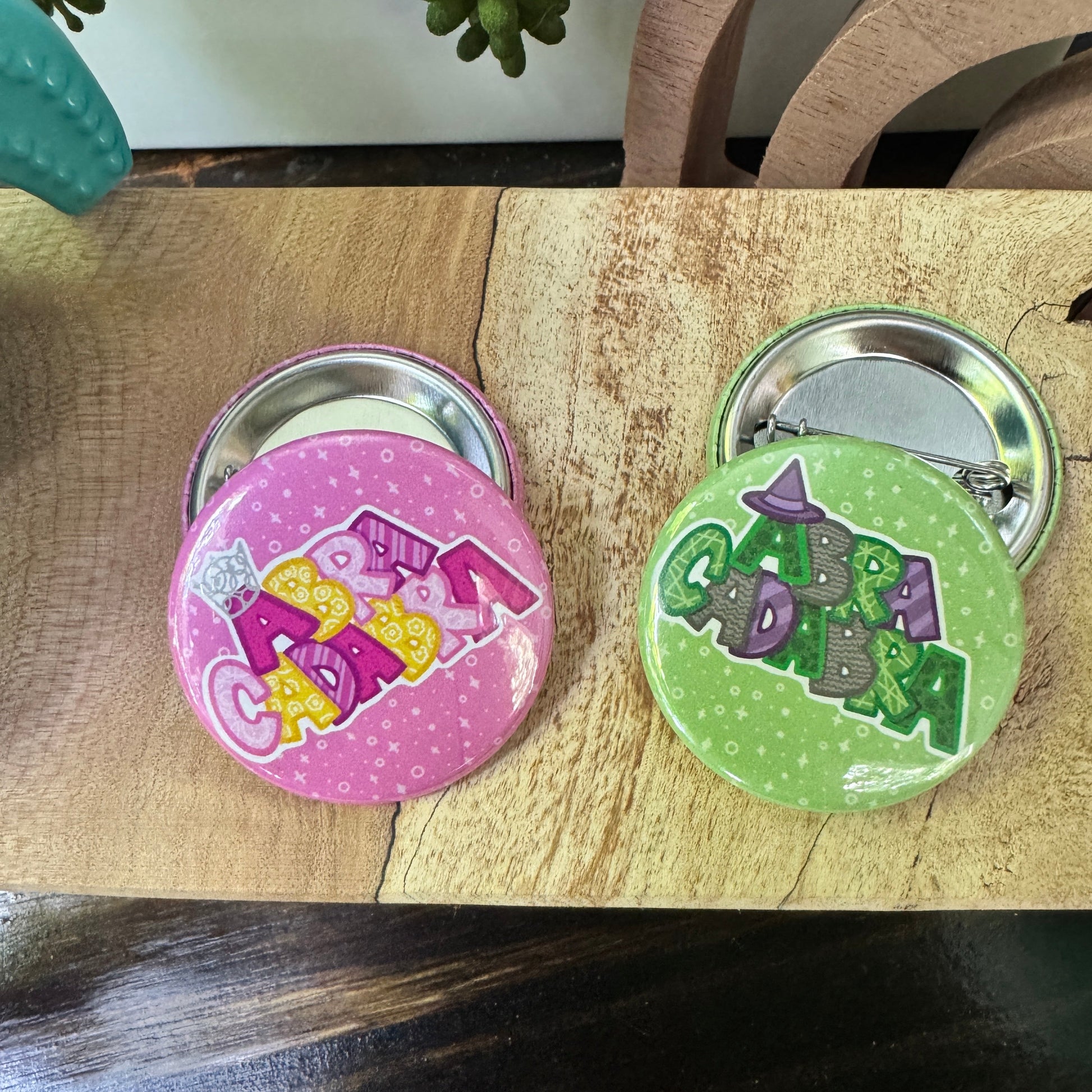 Two buttons, one green and one pink, on a wooden surface. Both say Abracadabra, one with a witches hat, one with a sparkl tiara.
